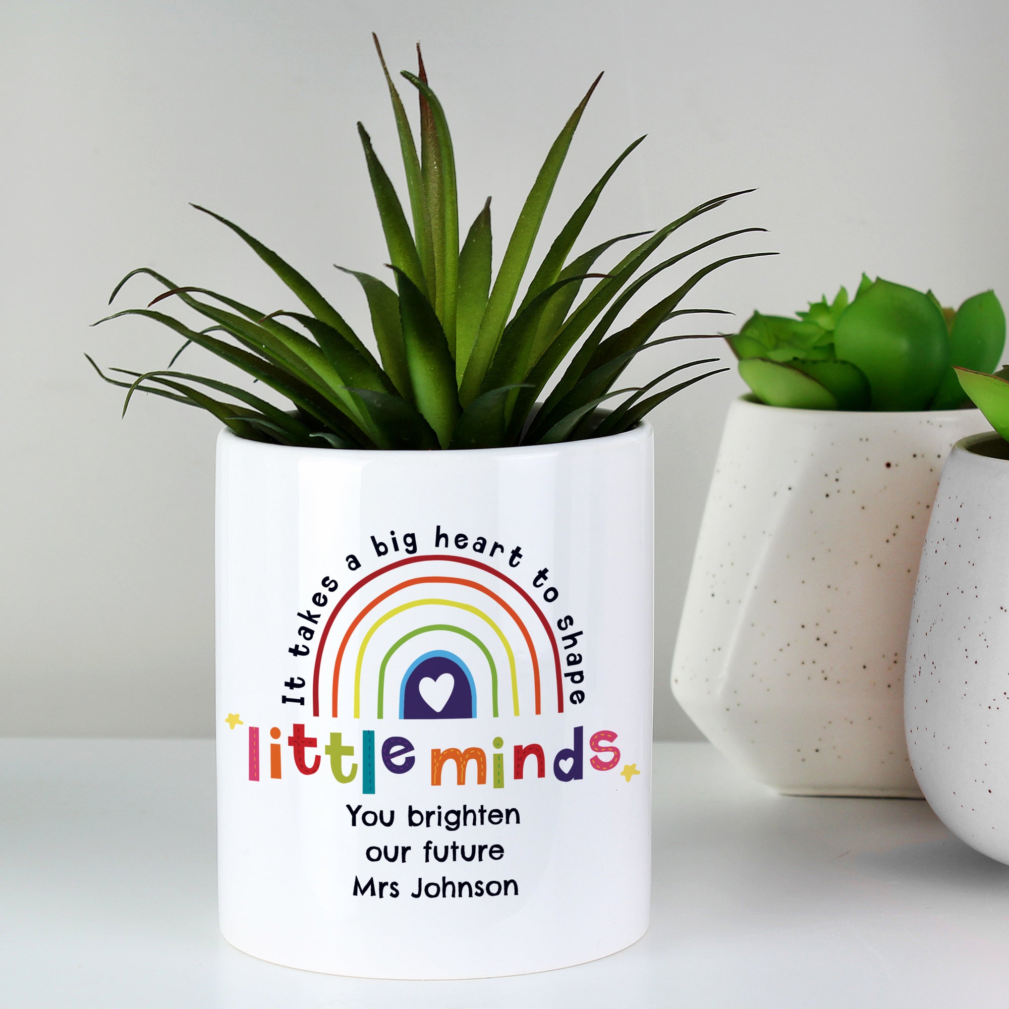 Image of a white ceramic pot with the text 'it takes a big heart to shape little minds' printed around a multicoloured rainbow. Directly below the pot can be personalised with your own text over up to 3 lines as a thank you gift for a teacher or teaching assistant.