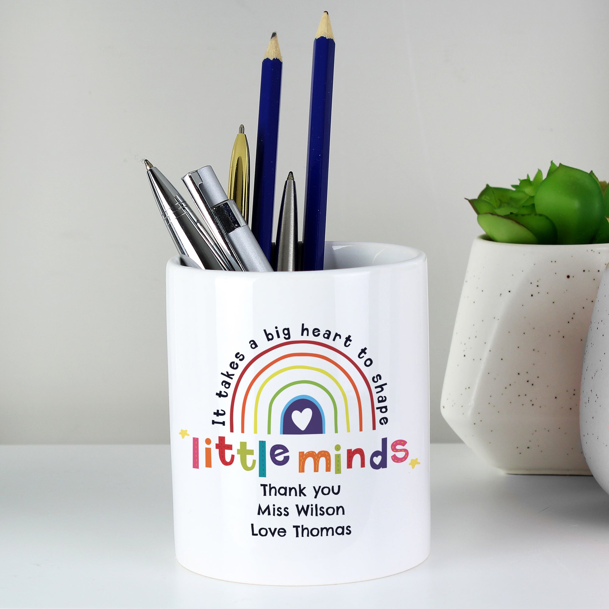 Image of a white ceramic pot with the text 'it takes a big heart to shape little minds' printed around a multicoloured rainbow. Directly below the pot can be personalised with your own text over up to 3 lines as a thank you gift for a teacher or teaching assistant.