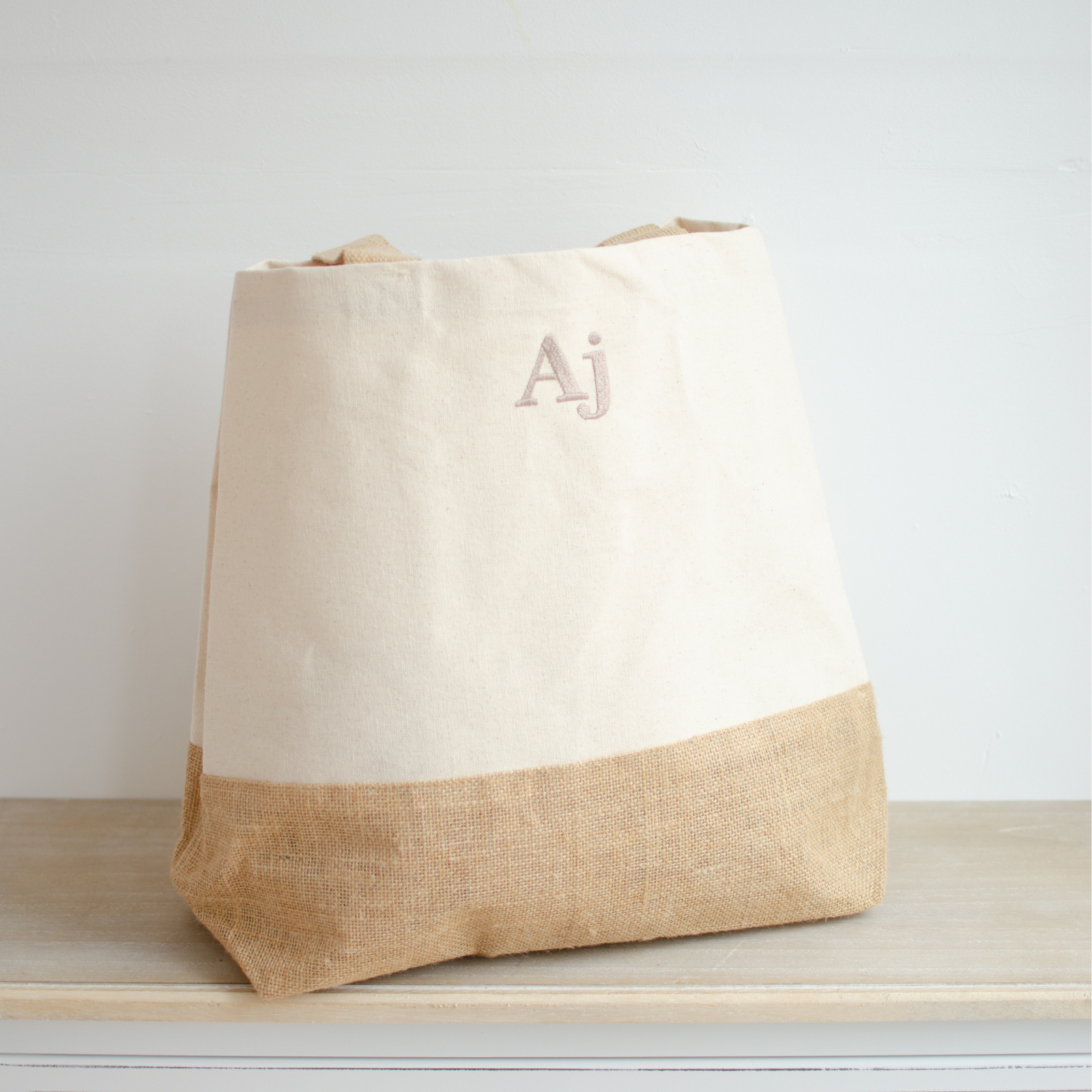 Image of a personalised neutral coloured bag that has been personalised. The bag has a jute base which is coloured a light brown and the main body of the bag is a neutral coloured canvas. The bag has been personalised in colour coordinated name. The bag has two shoulder straps that are the same colour as the jute base.