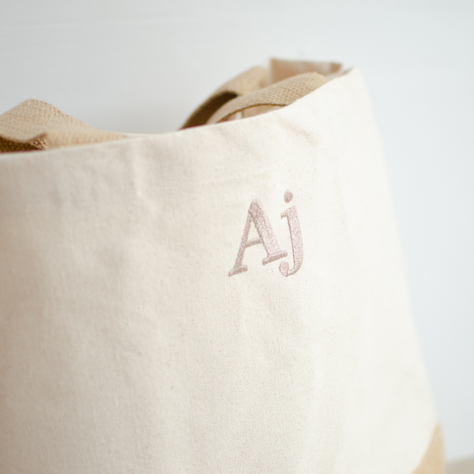 Image of a personalised neutral coloured bag that has been personalised. The bag has a jute base which is coloured a light brown and the main body of the bag is a neutral coloured canvas. The bag has been personalised in colour coordinated name. The bag has two shoulder straps that are the same colour as the jute base.