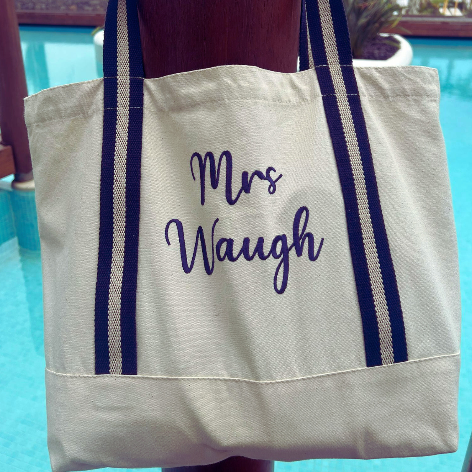 Image of a natural coloured tote bag with navy blue straps that has been personalised with a name embroidered in navy thread. The bag is available with four different strap colours, amber, red, navy and sage green and will be personalised in a matching thread colour.