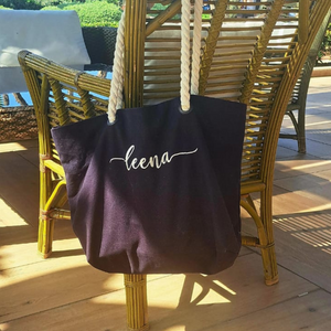 Image of a navy tote bag with natural coloured rope handles hanging on the back of a chair in the sunshine. The bag is personalised with a name which has been embroidered in a natural coloured embroidery thread in your choice of fonts.