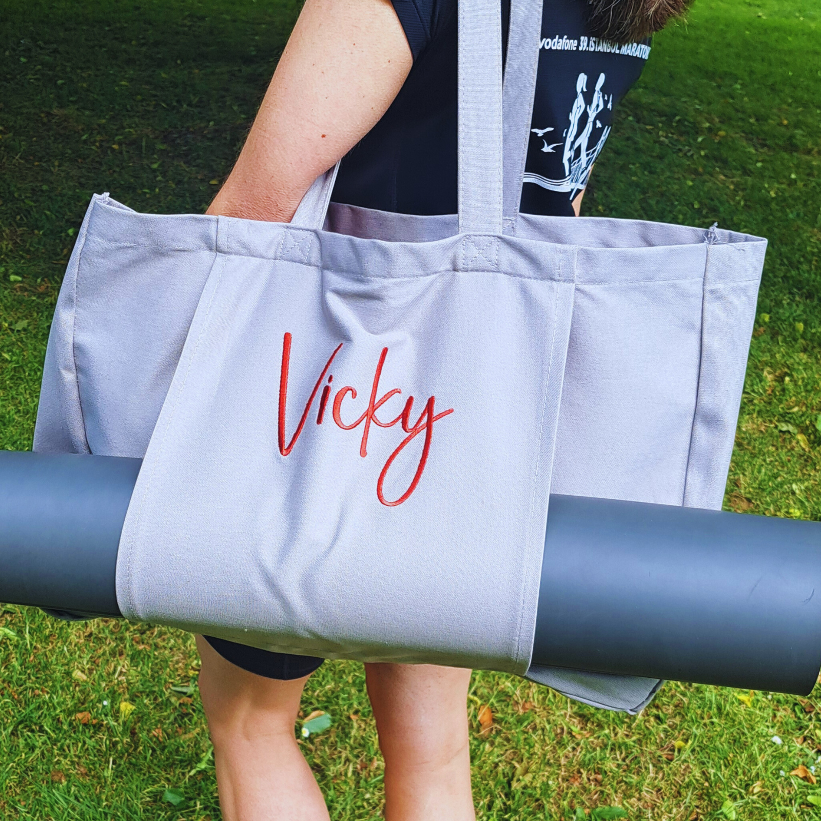Image of a grey yoga mat bag being carried on a shoulder. The bag has a large internal space for all of your yoga essentials and the front of the bag has a sleeve to hold your rolled up yoga mat. The front of the bag can be personalised wtih a name in a font and colour of your choice.