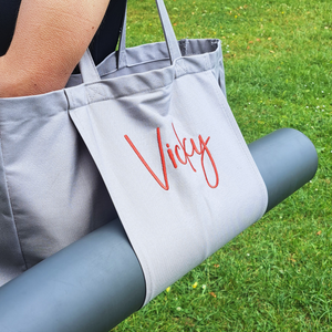 Image of a grey yoga mat bag being carried on a shoulder. The bag has a large internal space for all of your yoga essentials and the front of the bag has a sleeve to hold your rolled up yoga mat. The front of the bag can be personalised wtih a name in a font and colour of your choice.